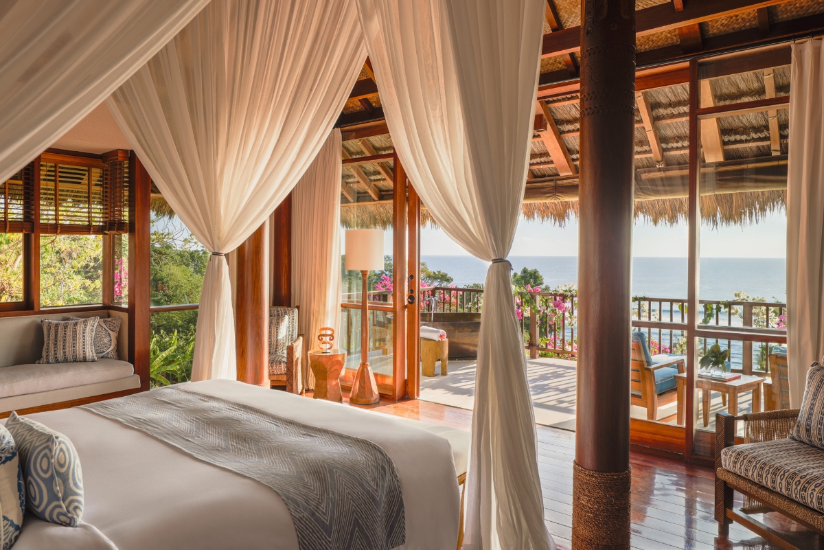 The image shows a tropical-style bedroom featuring a four-poster bed with flowing white curtains and a thatched palm roof ceiling. Large open doorways lead to a wooden terrace with ocean views, and the room includes wicker furniture and polished wooden floors.