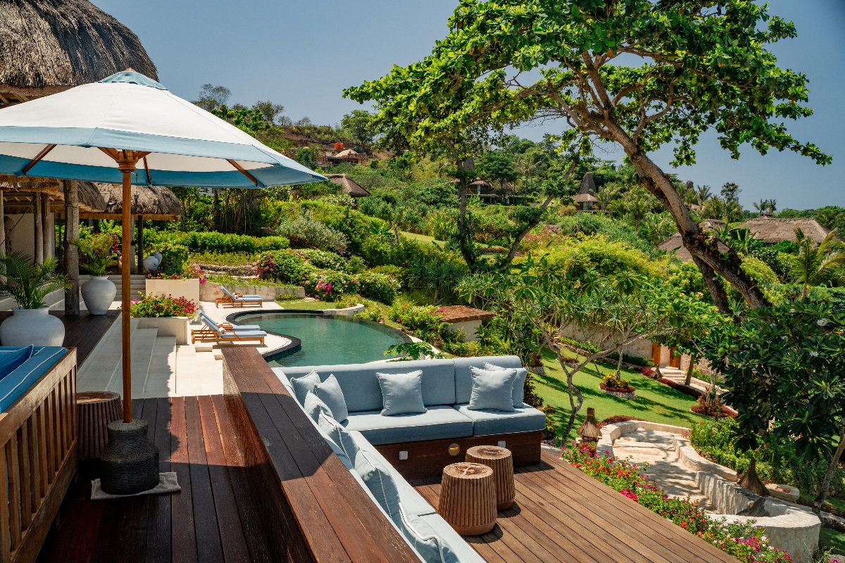 A luxury outdoor terrace features a wooden deck with a white sectional sofa, wooden side tables, and a large white umbrella, overlooking a swimming pool surrounded by lush tropical landscaping. The elevated deck provides views of rolling green hills dotted with traditional thatched-roof buildings and dense vegetation under a clear sky.