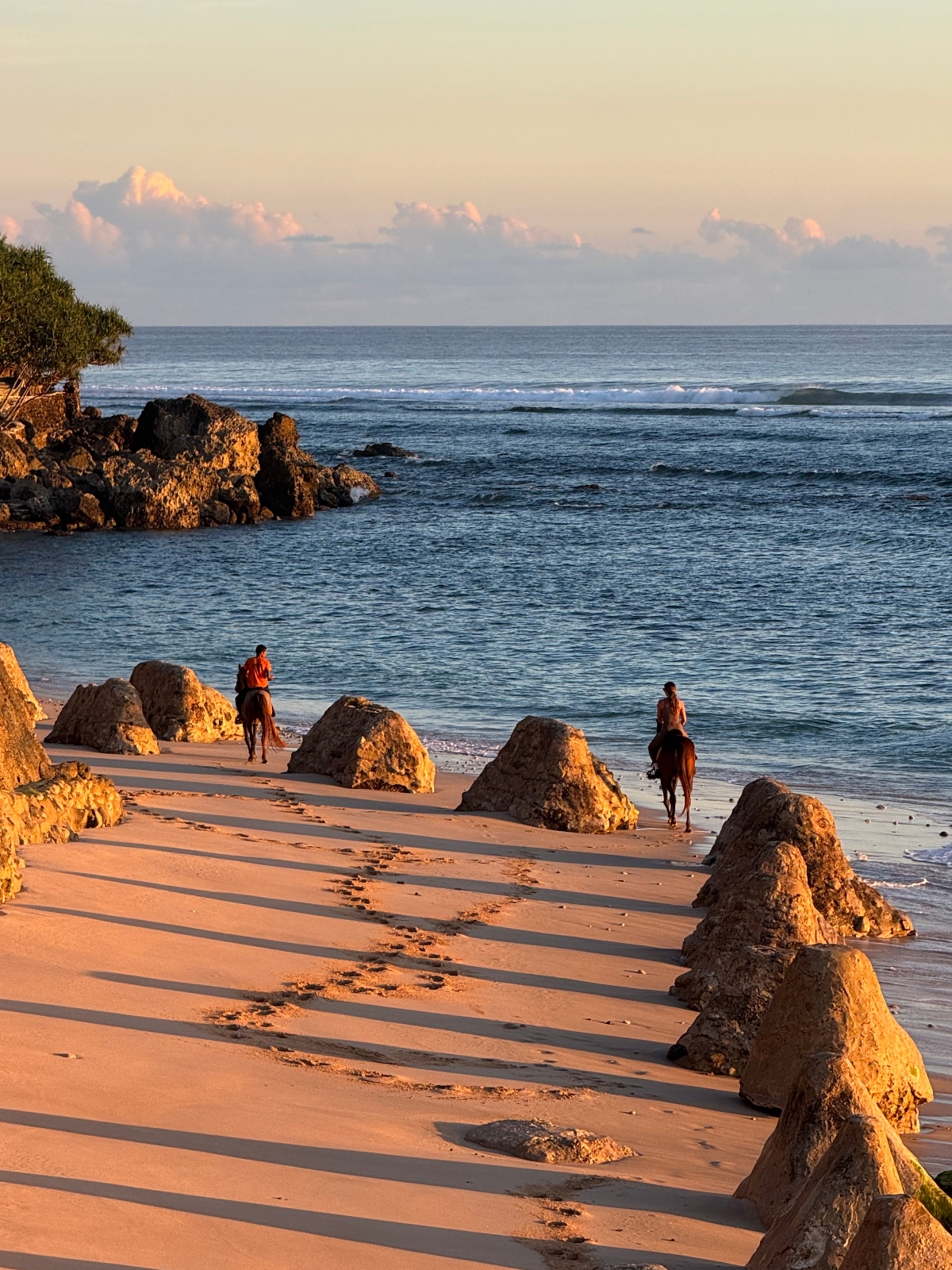 A beach scene during golden hour shows two horseback riders on sandy shores dotted with large rock formations that cast long shadows across the sand. The ocean stretches to the horizon with gentle waves rolling in, while rocky cliffs are visible on the left side under a cloudy sky with warm lighting.