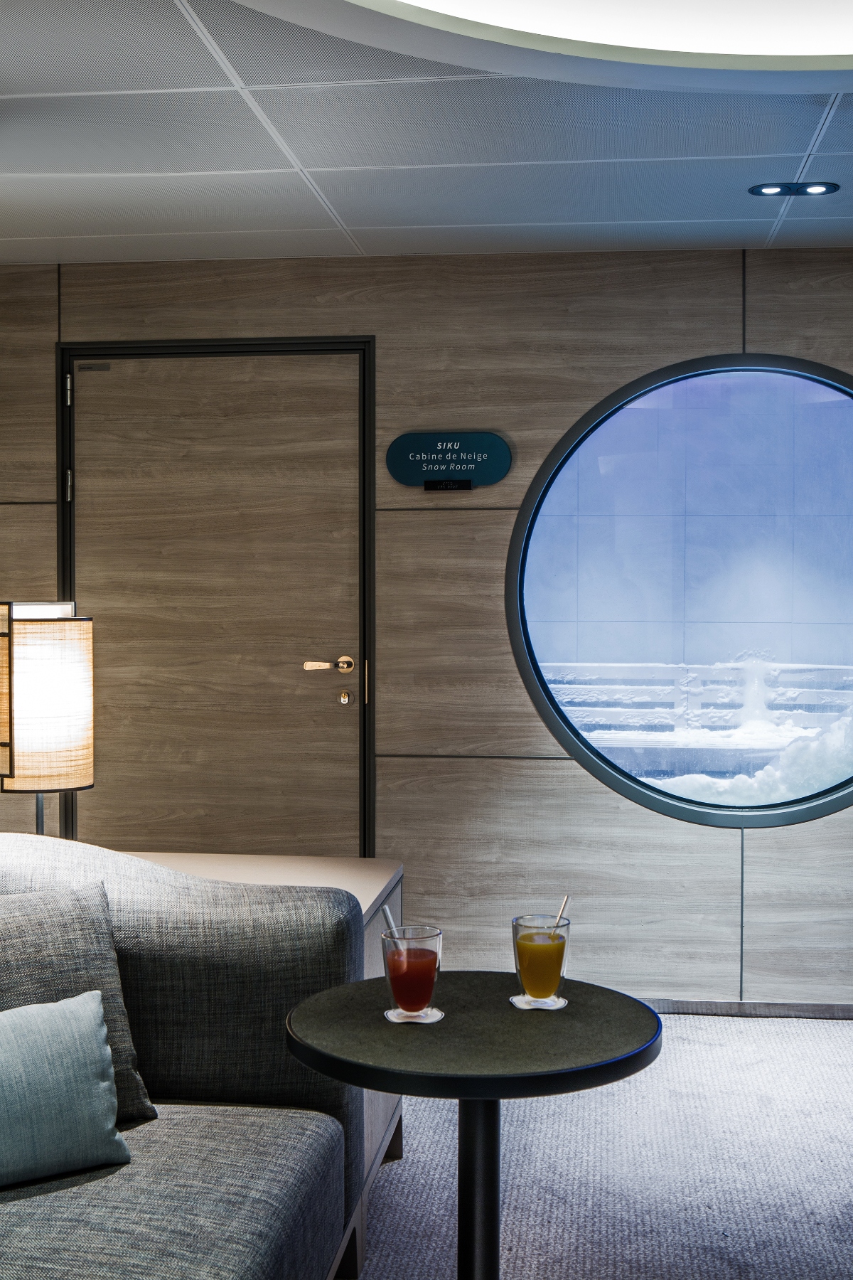 A modern ship cabin interior features a large circular porthole window revealing an icy space beyond. The room includes contemporary furnishings with gray upholstered seating, a small round black table holding two beverages, and wood-grain wall paneling. A door marked "SIKU Cabine de Neige Snow Room" and a floor lamp complete the luxurious cabin setting.