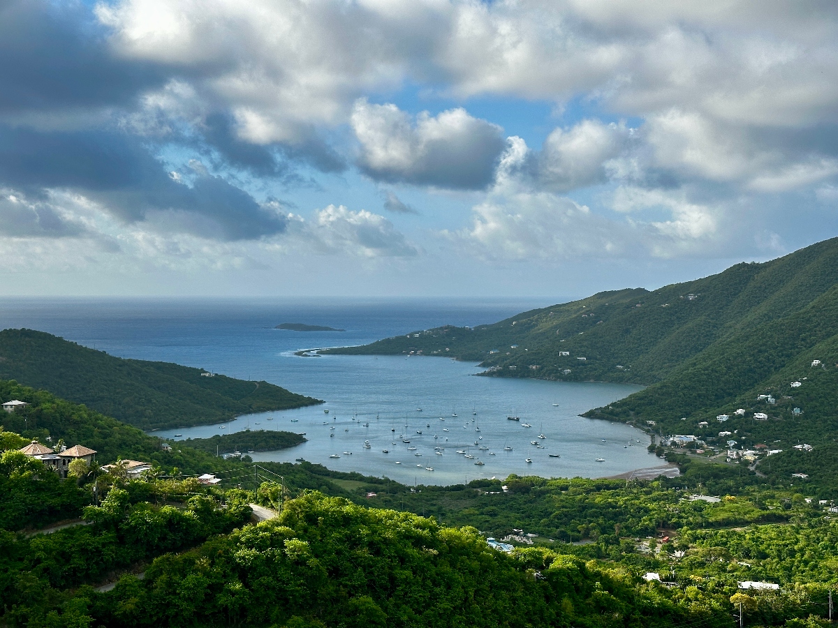 This aerial view shows a large, protected bay filled with dozens of small boats at anchor, surrounded by lush, green forested hills dotted with white residential buildings. The bay opens to deeper blue waters in the distance, while the verdant landscape rises steeply from the shoreline on multiple sides. Puffy white clouds drift across a partly cloudy blue sky above this tropical or subtropical coastal setting.