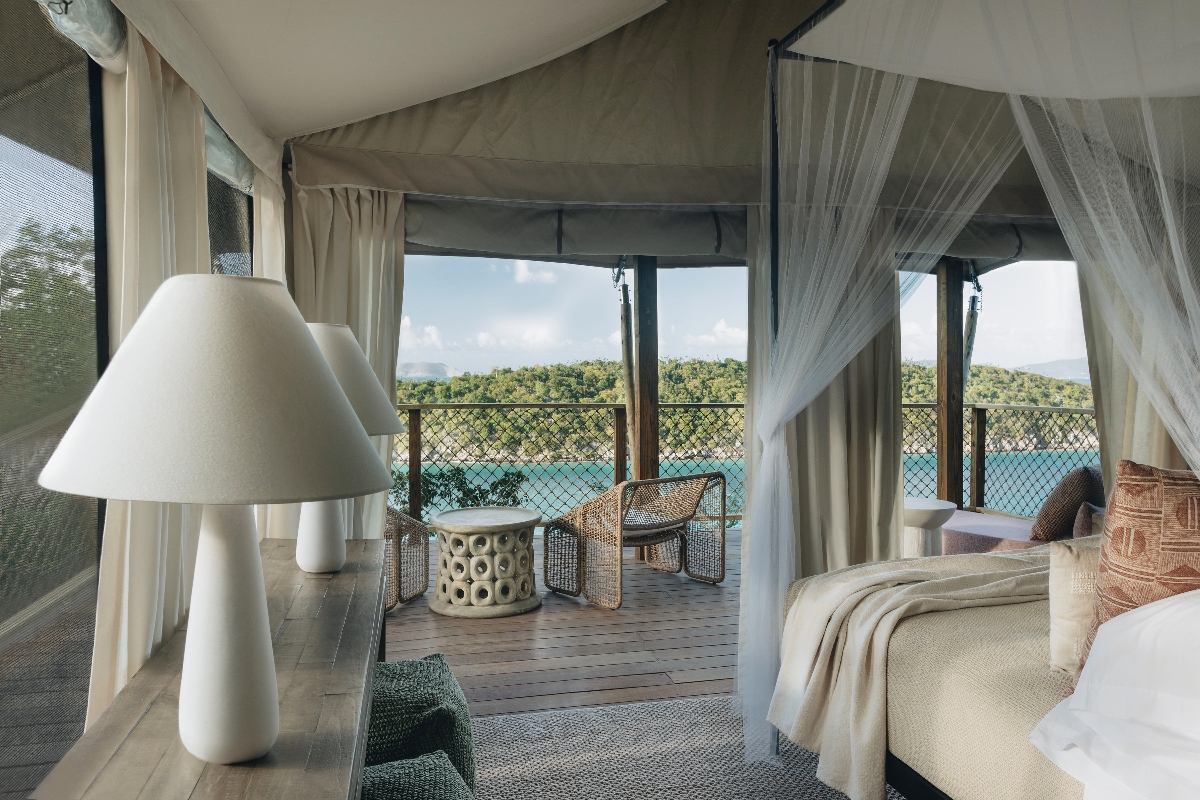 A glamping tent interior features a bed with white linens and neutral-toned pillows, with large glass doors opening onto a wooden deck terrace that overlooks the ocean and forested hills. The space includes a white sculptural table lamp, flowing white curtains, and wicker furniture on the outdoor terrace area.