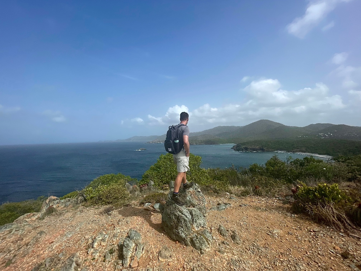 A person wearing a backpack stands on rocky terrain overlooking a coastal landscape with blue water and forested hills. The viewpoint offers a panoramic view of a bay or inlet surrounded by green mountainous terrain under a partly cloudy blue sky.