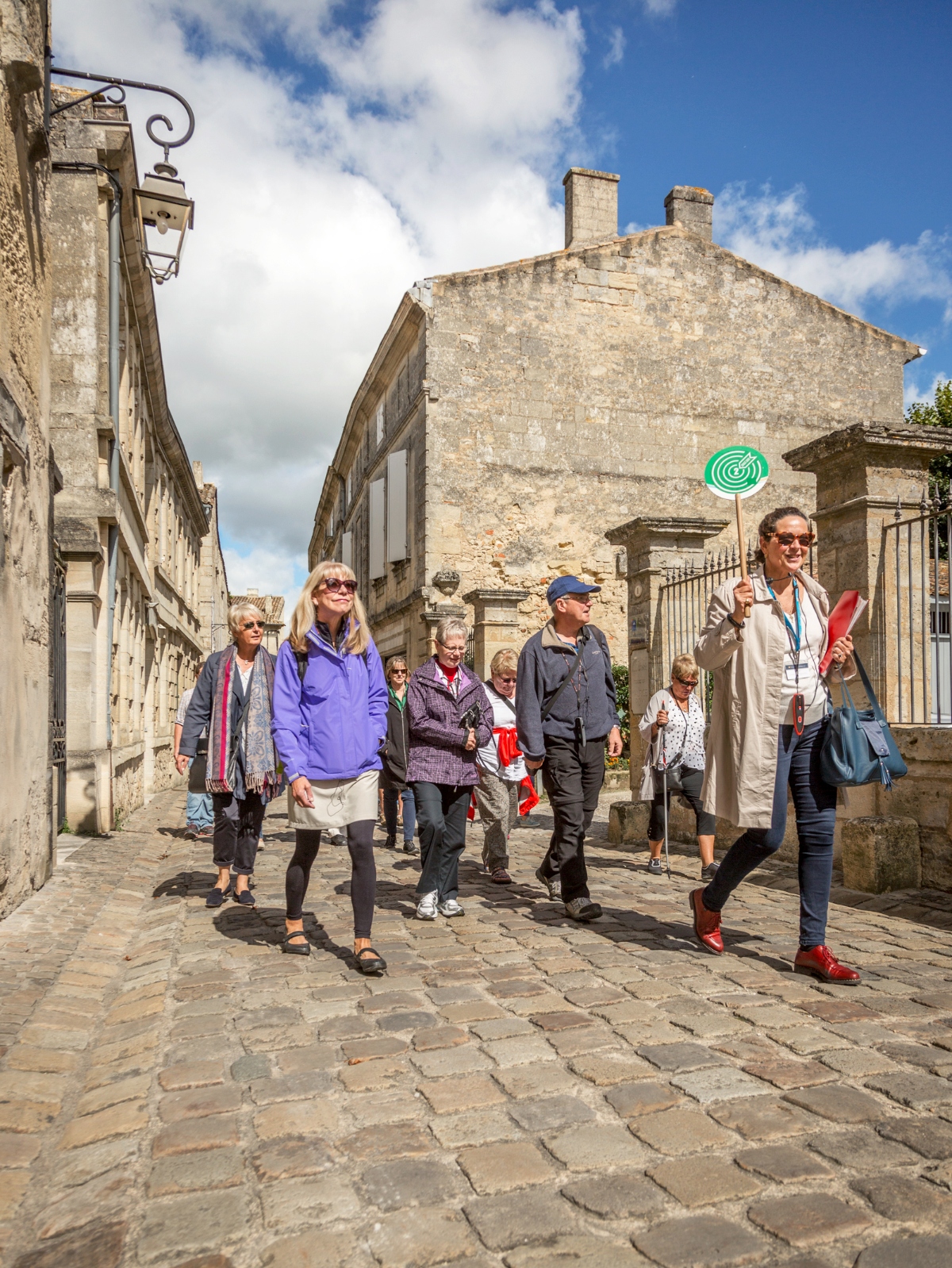 A group of approximately ten older adults walks along a cobblestone street in what appears to be a historic European town, with one person wearing a lanyard and holding red materials who appears to be leading the group. The scene shows traditional limestone buildings with weathered stone facades, a wrought iron wall-mounted lantern, and a partly cloudy blue sky overhead.