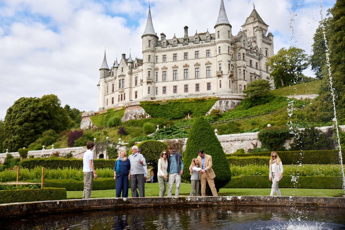 A magnificent white stone castle with multiple conical towers and turrets sits atop a hillside, surrounded by lush green terraced gardens and mature trees. In the foreground, a group of approximately eight tourists stands on manicured lawns near a water fountain, with the visitors wearing casual clothing and appearing to be on a sightseeing tour. The formal gardens feature precisely trimmed hedges, topiary shapes, and stone balustrades that create elegant terraced levels leading up to the imposing castle structure.