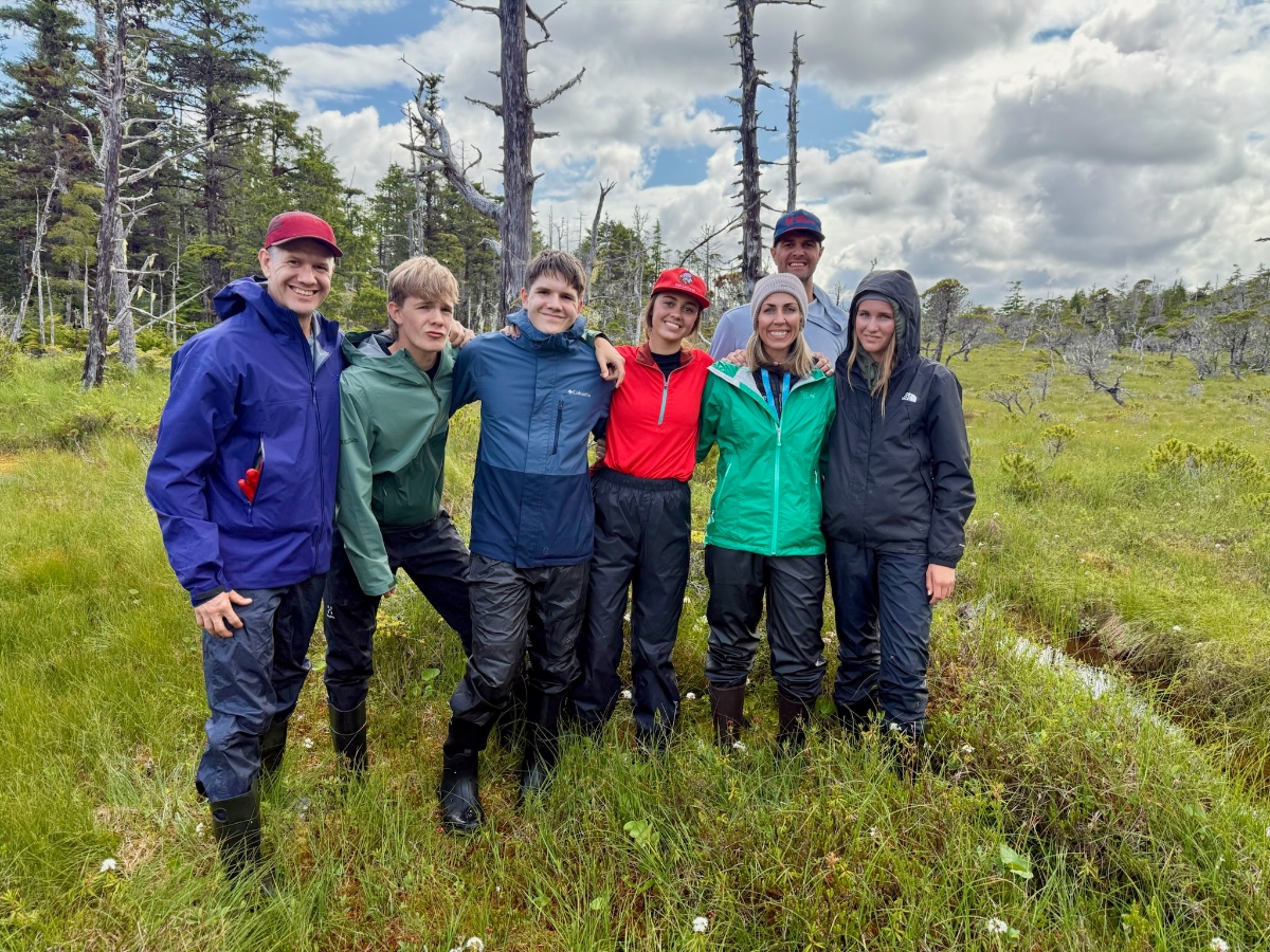A group of seven people wearing outdoor gear including rain jackets, boots, and caps pose together in a marshy wetland area with tall grass. The landscape behind them features a mix of bare, dead trees and living evergreens under a cloudy sky. The group members are dressed in various colored jackets including blue, green, red, and black, suggesting they are prepared for wet or changeable weather conditions.