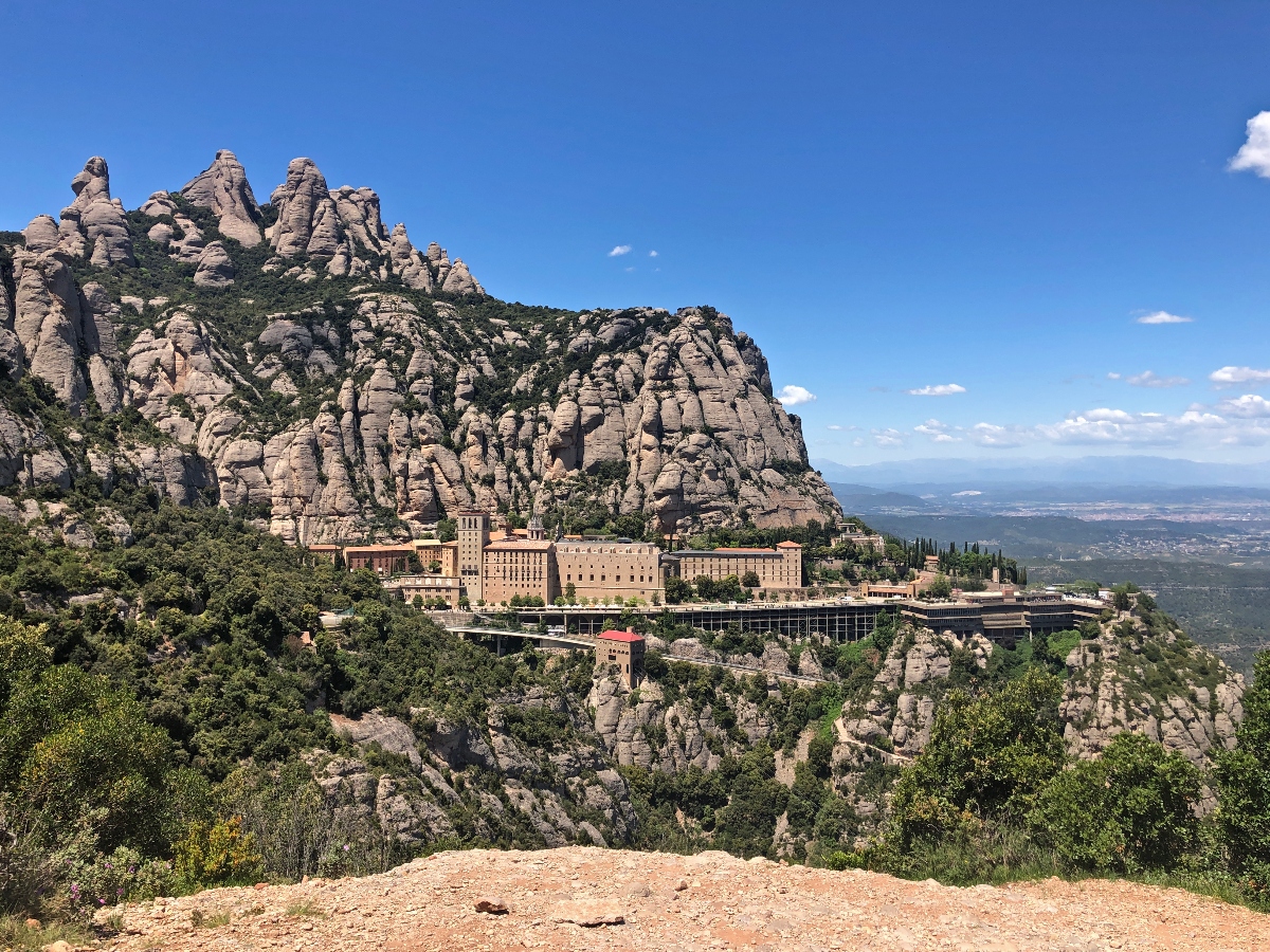 A large monastery complex with traditional stone buildings is nestled against dramatic, towering rock formations consisting of distinctive rounded stone pillars and spires. The religious buildings appear to include both historic structures and modern facilities, with what appears to be cable car infrastructure visible among the buildings. The scene is set against a clear blue sky with the rocky mountain formations rising steeply above the complex, while a verdant valley landscape extends into the distance below.