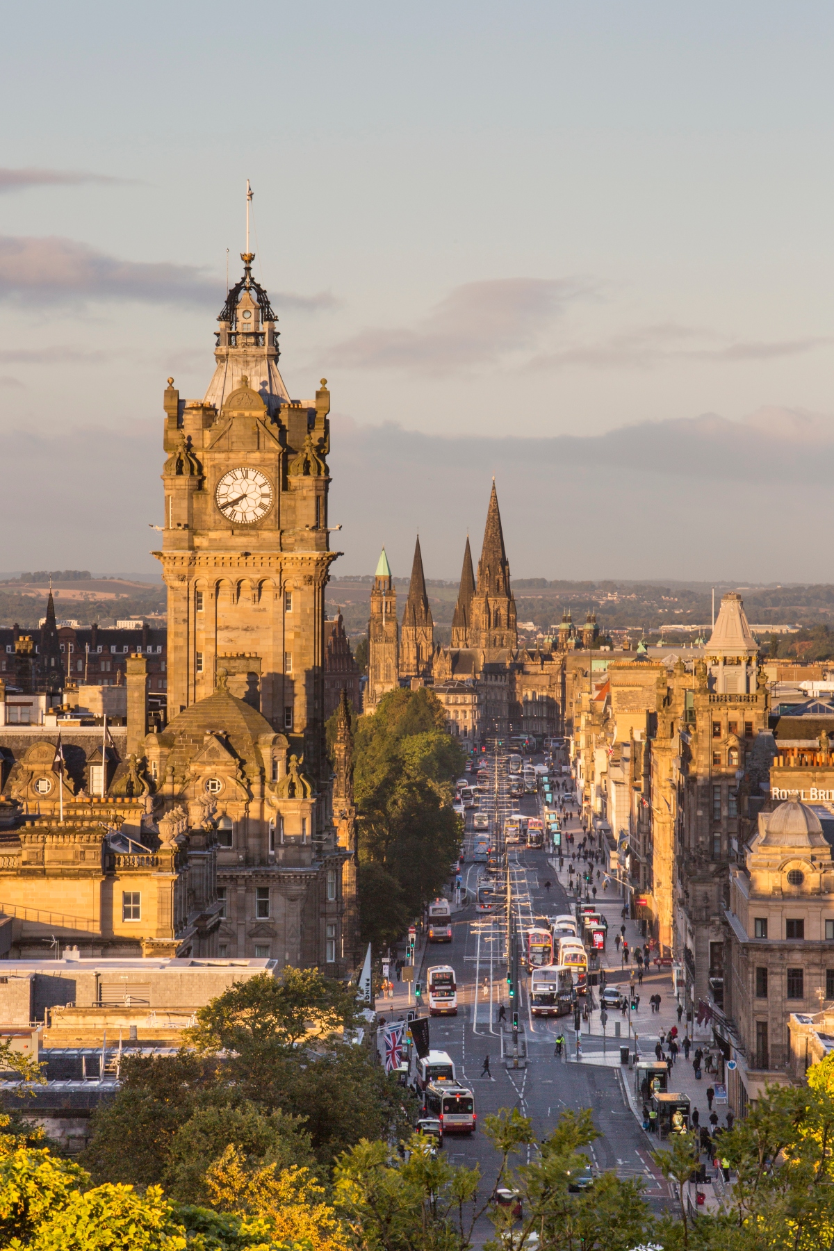 This elevated view of Edinburgh shows a prominent clock tower in the foreground with ornate Victorian architecture, while Gothic church spires are visible in the background across the cityscape. The busy street below features red buses, trams, and pedestrians moving along what appears to be a main thoroughfare lined with traditional sandstone buildings. The scene is captured during golden hour lighting, which illuminates the warm-toned stone architecture and creates long shadows across the urban landscape.