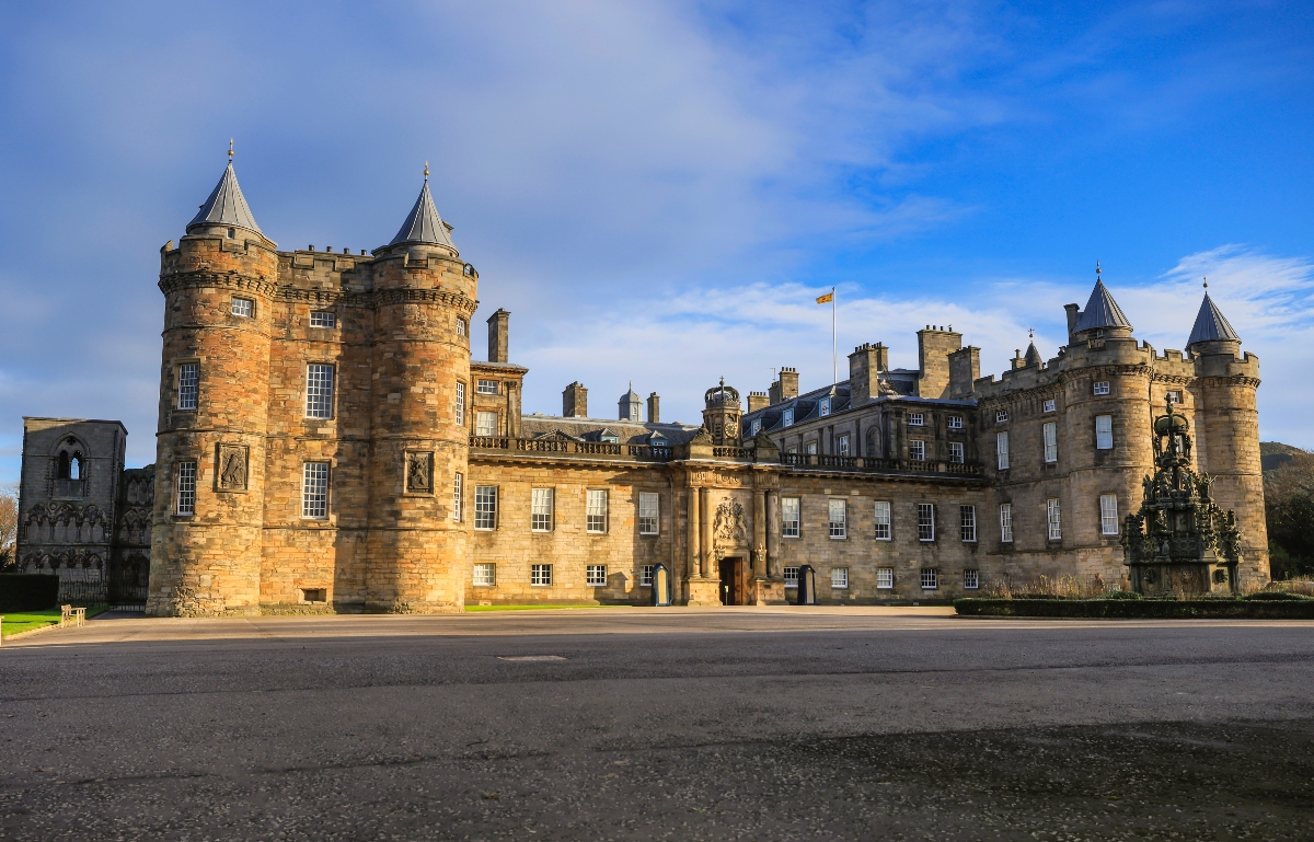 This image shows a large sandstone castle complex featuring multiple round towers with conical roofs and an adjoining multi-story palace building with numerous windows. The structure displays typical medieval Scottish architecture with warm golden stone, and a flag can be seen flying from one of the buildings. The castle sits beside a paved area or road, with some ruined structures visible on the left side, all photographed under a partly cloudy blue sky.