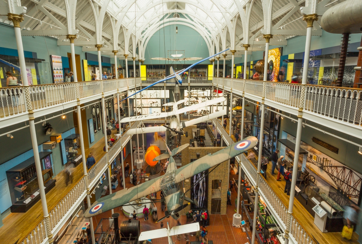 This image shows the interior of a large multi-story museum with Victorian-era white cast iron architecture, featuring ornate columns, arches, and decorative balustrades across several levels. Multiple aircraft are suspended from the ceiling throughout the spacious atrium, including what appears to be military aircraft with RAF roundels and other vintage planes displayed at various heights. Visitors can be seen exploring the exhibits on different floors connected by walkways and galleries, with colorful display panels and exhibits visible along the perimeter walls.