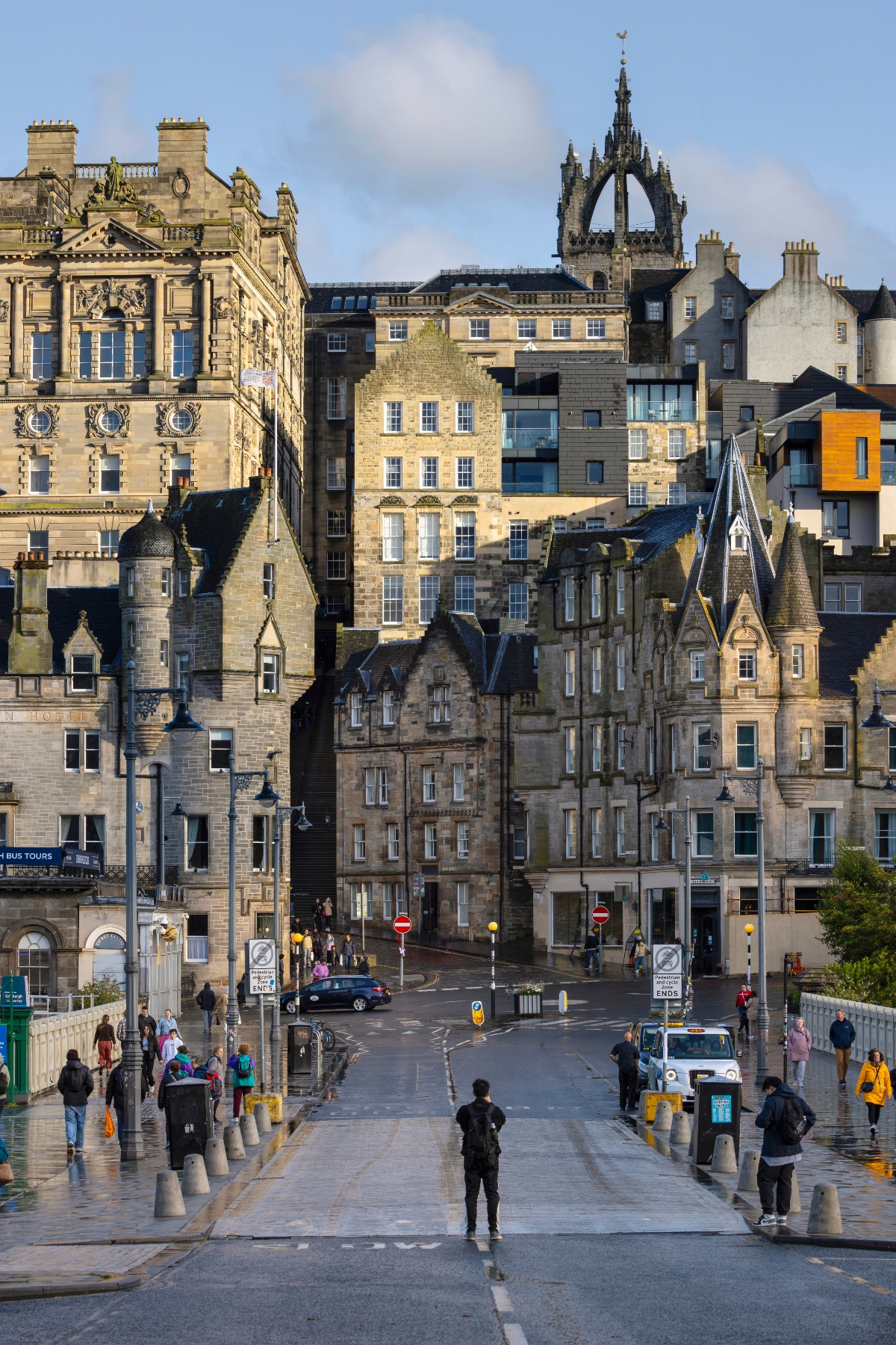 This image shows a historic city street lined with multi-story sandstone buildings in traditional Scottish architectural style, with a prominent Gothic church spire visible in the background. The scene captures pedestrians walking along the street and sidewalks in what appears to be late afternoon lighting that casts a warm glow on the stone facades. The view is taken from an elevated perspective looking down the street, showing a mix of historic architecture with some modern additions integrated among the older buildings.