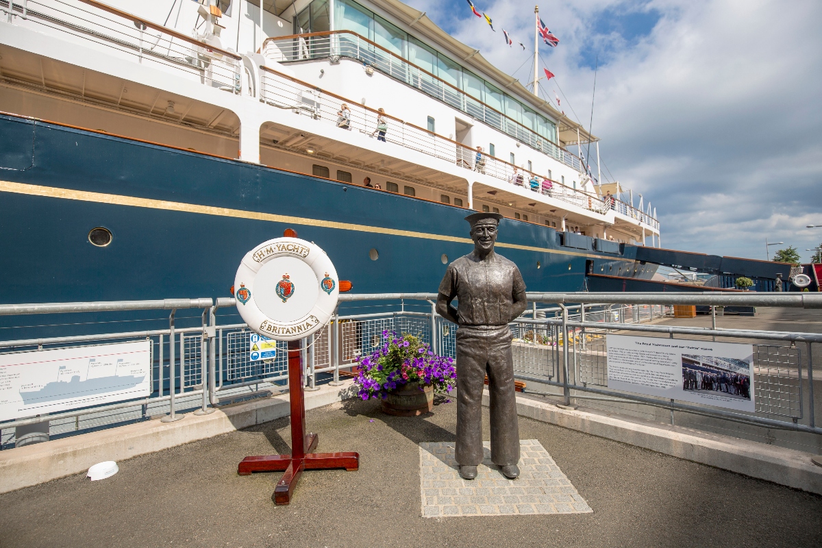 This image shows a bronze statue of a uniformed figure standing on a platform next to a large ship with a blue hull and white superstructure docked at a pier. A white life ring labeled "HMY YACHT BRITANNIA" is displayed on a red stand beside the statue, along with purple flowering plants and informational displays behind metal railings. The scene is captured under a partly cloudy sky with flags visible flying from the ship's rigging.