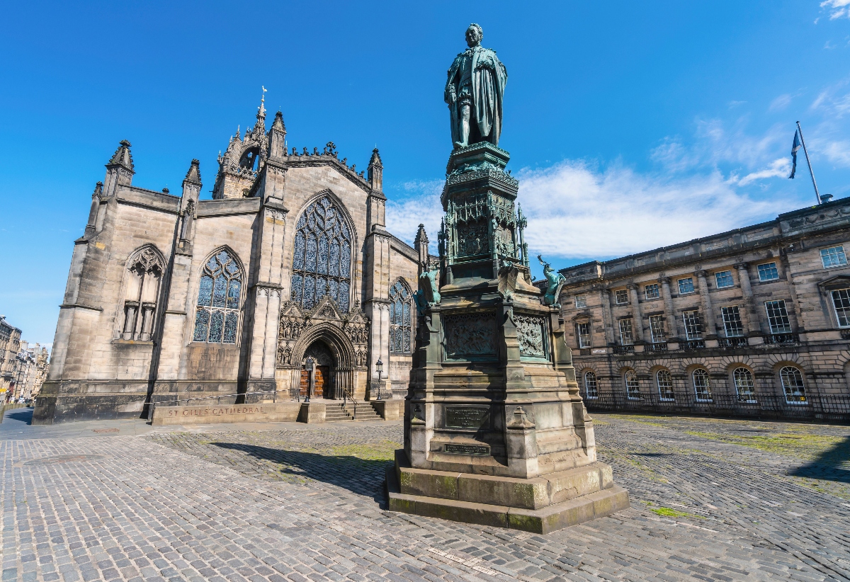 This image shows a cobblestone plaza featuring a prominent bronze statue of a robed figure on an ornate stone pedestal, positioned in front of St Giles Cathedral, a Gothic stone church with large arched windows and decorative spires. Classical sandstone buildings line the right side of the square, and a blue flag can be seen flying from one of the structures. The scene is captured under a bright blue sky with scattered white clouds, creating strong shadows across the paved courtyard.