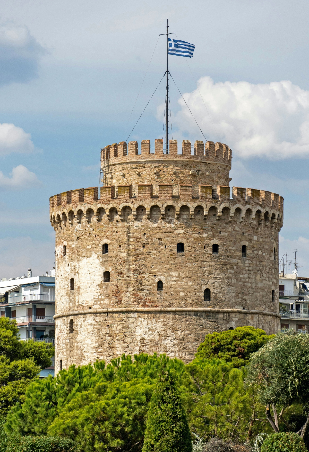 This image shows a large, circular stone tower with distinctive crenellated walls and multiple tiers, featuring small rectangular windows throughout its weathered masonry construction. A Greek flag flies from a tall flagpole mounted at the tower's summit, while the structure appears to be surrounded by lush green vegetation in the foreground. Modern residential buildings are visible in the background, contrasting with the ancient fortress-like architecture of the prominent cylindrical tower.