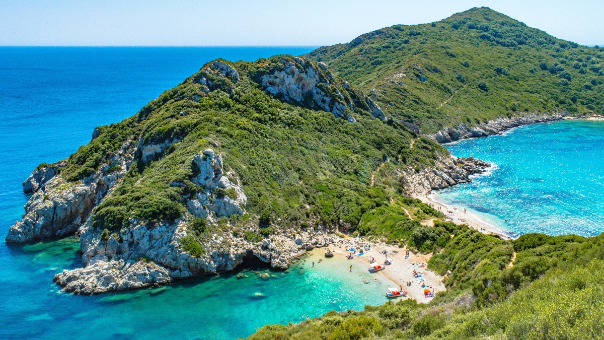 This aerial view shows a small, secluded beach nestled between steep, forested hills and dramatic white limestone cliffs along a Mediterranean-style coastline. The sheltered cove features crystal-clear turquoise waters with visible beachgoers and colorful umbrellas or equipment scattered across the sandy shore. The surrounding landscape is characterized by dense green vegetation covering rolling hills that drop sharply into the brilliant blue sea, with rocky outcroppings creating natural barriers around the pristine bay.