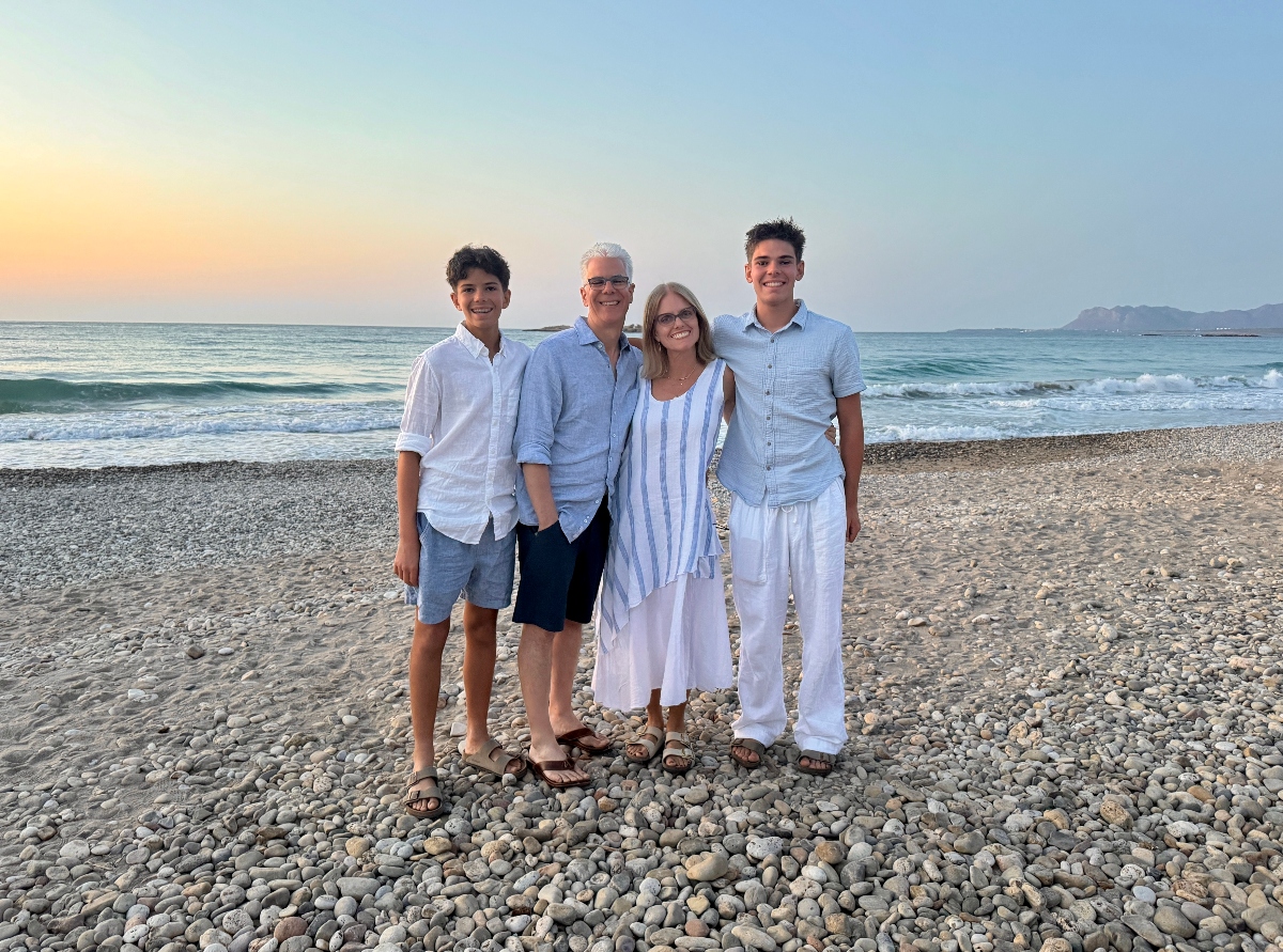 Four people stand together on a pebbled beach wearing coordinated light blue and white clothing, with ocean waves and a sunset sky visible behind them. The beach setting features a rocky shoreline with mountains visible in the distance across the water.