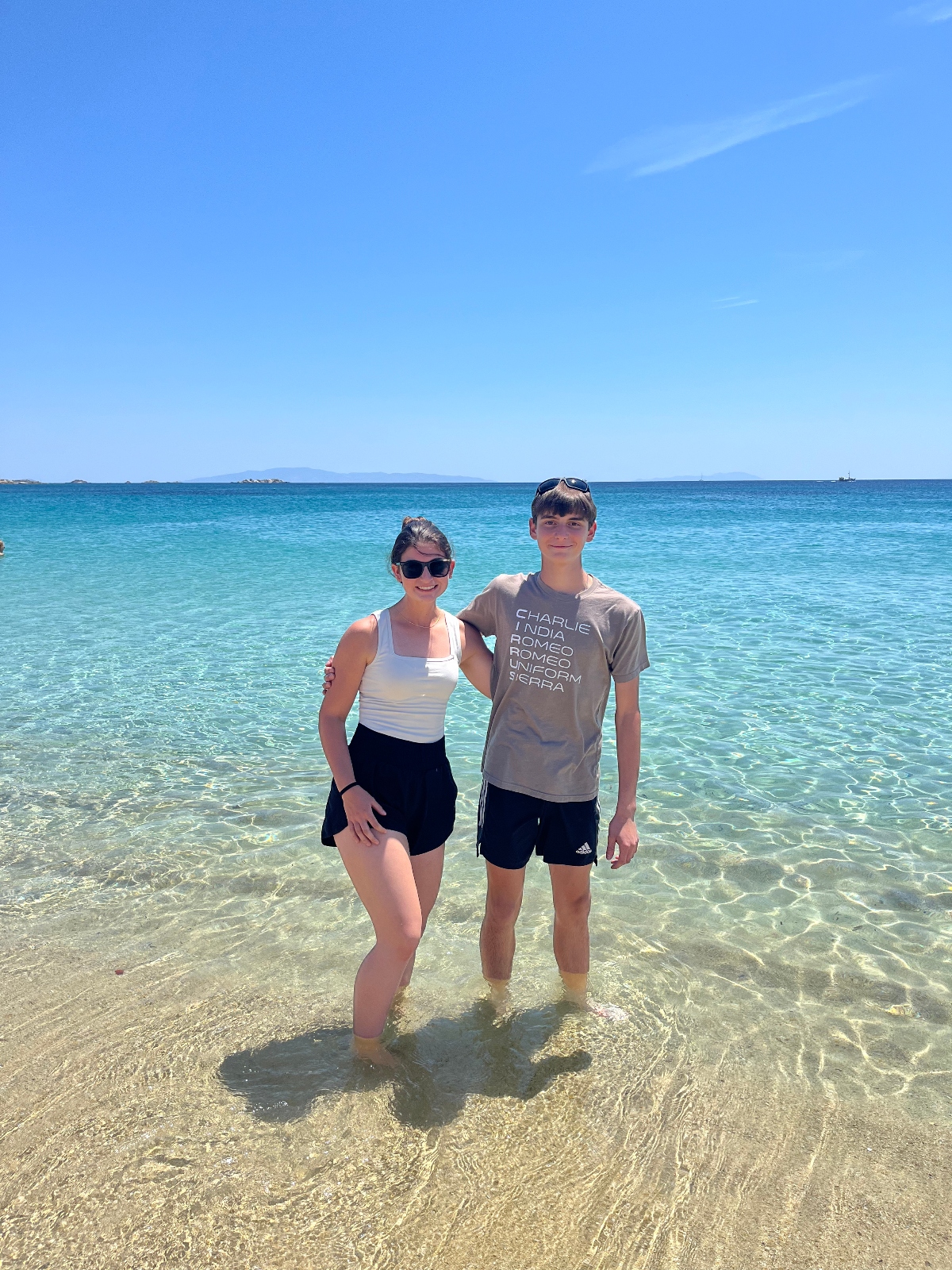 Two people stand together in shallow, crystal-clear turquoise water at a beach, with the person on the left wearing sunglasses and a white tank top, and the person on the right wearing a gray t-shirt. The scene shows a pristine Mediterranean setting with bright blue skies, distant islands on the horizon, and transparent water revealing the sandy bottom beneath their feet.