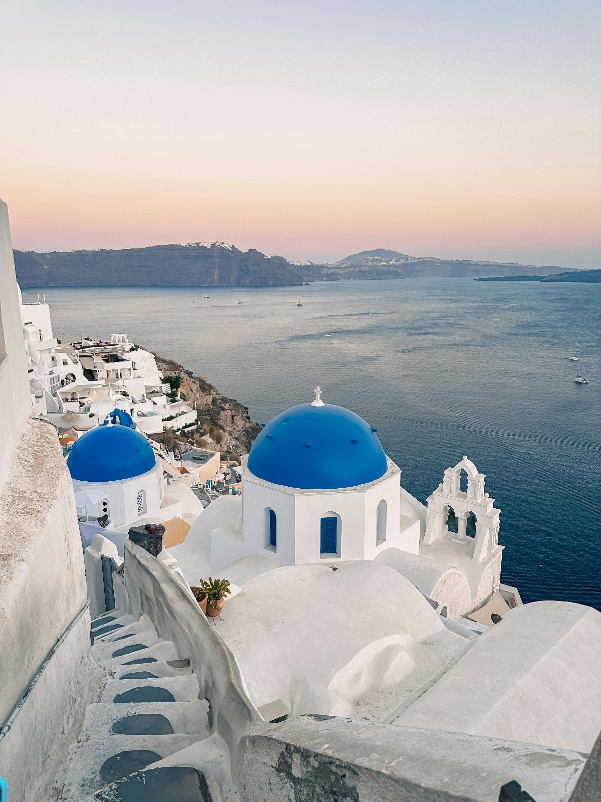 This image shows traditional white-washed buildings with distinctive bright blue domed roofs perched on a clifftop overlooking a calm sea. Stone steps curve downward through the terraced architecture, while the foreground features characteristic Mediterranean island buildings with their cubic forms and blue accents. In the background, distant islands are visible across the water under a soft, pastel sky suggesting either dawn or dusk lighting conditions.