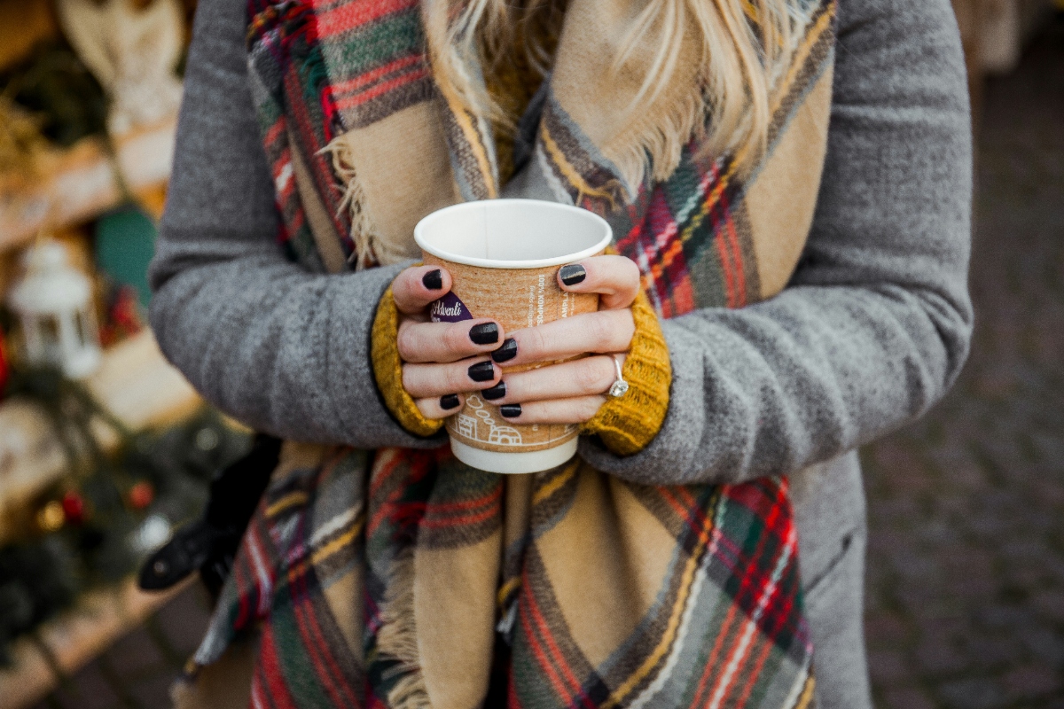 A person wearing a gray sweater and plaid scarf in tan, red, and green holds a disposable coffee cup with both hands in yellow mittens. The cup features a decorative pattern and the person's hands show dark nail polish. The blurred background suggests a Christmas market setting with out-of-focus lights and decorations.