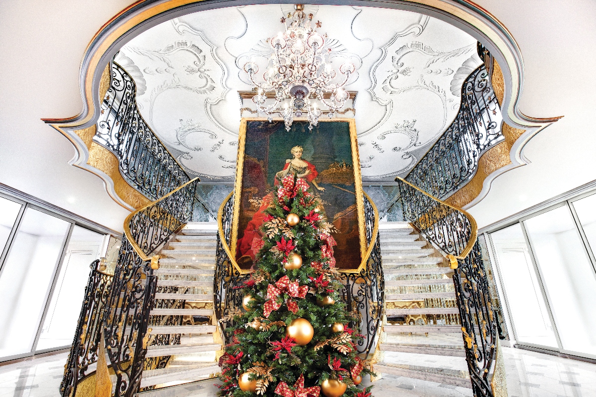 A grand symmetrical double staircase with white marble steps and ornate black wrought iron railings accented with gold trim, leading up to an elaborately decorated ceiling with baroque-style white plasterwork and a crystal chandelier. A decorated Christmas tree adorned with gold ornaments, red poinsettias, and festive greenery stands at the base of the stairs, positioned in front of a large gilt-framed classical portrait of a woman in period dress. The luxurious interior features polished surfaces and elegant architectural details throughout.
