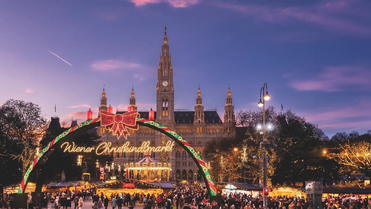 A festive Christmas market scene at dusk in Vienna, featuring an illuminated archway decorated with red and green lights and a large bow, with "Wiener Christkindlmarkt" written in glowing script. Behind the arch stands Vienna's neo-Gothic City Hall (Rathaus) with its prominent central tower and spire, beautifully lit against a pink and blue twilight sky. Crowds of visitors gather among illuminated market stalls and light-decorated trees in the square.