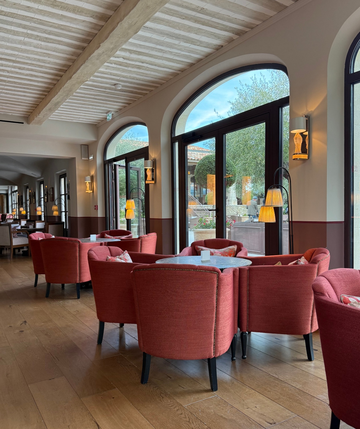 A hotel lounge or bar area features groupings of coral-colored upholstered armchairs with nailhead trim arranged around small round tables on wide-plank wood flooring. The room has exposed wooden ceiling beams with white panels between them, and large arched French doors with dark frames that open to a courtyard view with manicured topiary trees. Amber-shaded wall sconces and arc floor lamps provide warm lighting throughout the space, which has cream-colored walls with darker wainscoting at the lower portion.