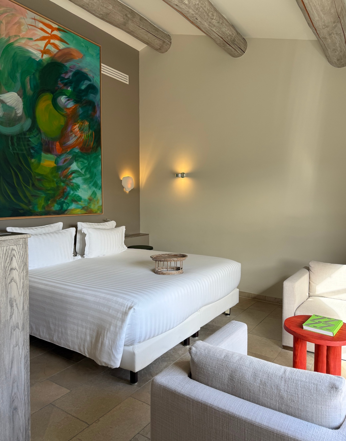 A modern hotel bedroom featuring exposed wooden ceiling beams and neutral beige walls with wall-mounted sconces. The room contains a bed dressed in white striped linens positioned against a wall displaying a large abstract painting in vibrant greens, oranges, and yellows. A seating area with gray upholstered furniture and a distinctive red round side table holding a green book occupies the foreground, with tile flooring throughout the space.
