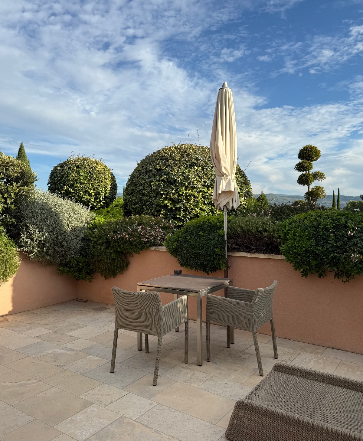 A private patio with beige stone tile flooring features wicker outdoor furniture including a dining table with four chairs and a closed cream-colored umbrella. The terrace is enclosed by terra cotta-colored walls topped with manicured spherical topiaries and lush greenery that creates a natural privacy screen. Beyond the landscaping, cypress trees and rolling hills are visible under a partly cloudy blue sky.