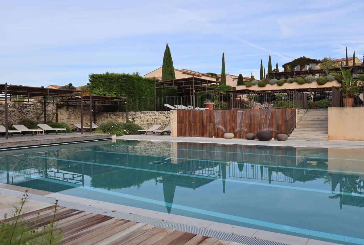 A large outdoor swimming pool with still turquoise water reflects the surrounding architecture and cypress trees. The pool area features wooden deck sections, white sun loungers positioned under pergolas on the left, and a distinctive weathered metal panel wall with spherical decorative stones serving as a water feature on the right. Stone walls, terraced levels, and Mediterranean-style buildings with cypress trees are visible in the background under a light blue sky.