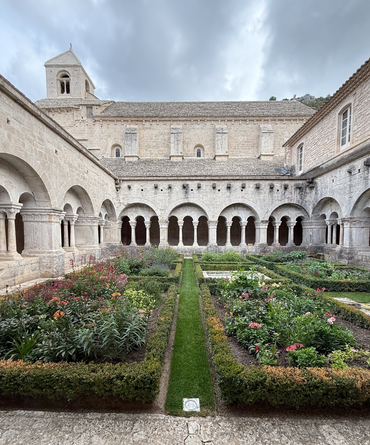 A medieval monastery cloister features a square courtyard surrounded by Romanesque stone arcades with rounded arches supported by columns. The interior courtyard contains a formal garden with symmetrical planted beds divided by a central grass pathway, featuring flowering plants and low hedges arranged in geometric patterns. A bell tower with arched openings rises above the stone buildings under an overcast sky.