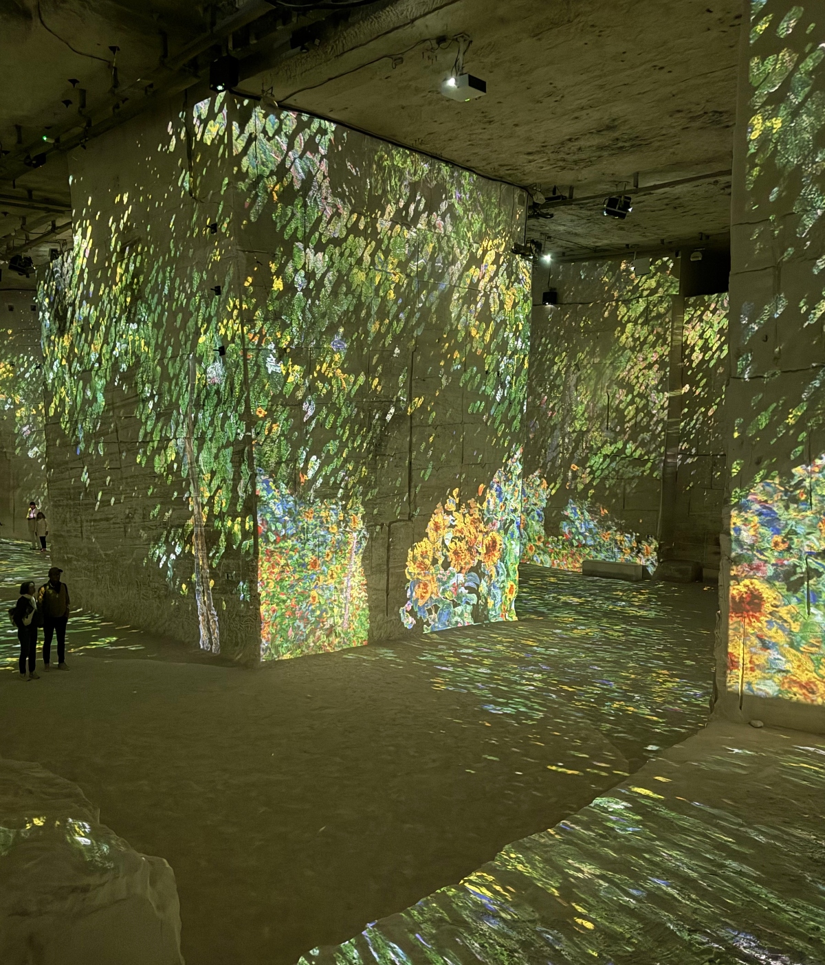 An immersive projection installation in a large carved stone space displays colorful artwork across walls, pillars, ceiling, and floor, featuring imagery with vibrant greens, yellows, blues, and oranges that appears to depict natural scenes and floral elements. The projections wrap around the architectural features of the rough stone interior, creating an enveloping visual environment. Silhouettes of visitors can be seen on the left side of the expansive illuminated gallery space.