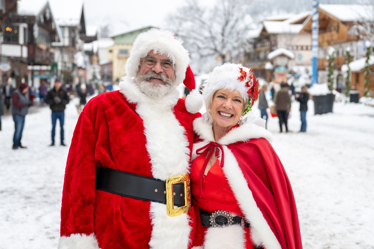 Two people dressed as Santa Claus and Mrs. Claus pose together in a snowy town square, with Santa wearing a traditional red suit with white fur trim and black belt, and Mrs. Claus wearing a red cape with white fur trim and a decorative headpiece. Behind them are Bavarian-style buildings with snow-covered roofs and crowds of people walking through the winter scene.