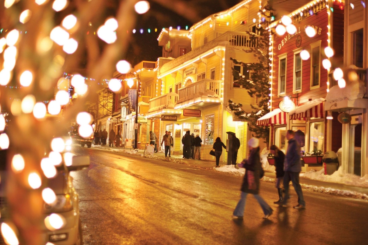 A historic main street at night features colorful two and three-story buildings with Victorian-era architecture illuminated by string lights, with snow-covered ground and pedestrians walking along the street. Bokeh lights from a decorated tree in the foreground create soft, glowing orbs that frame the view of the lit storefronts and snow-dusted evergreen trees.