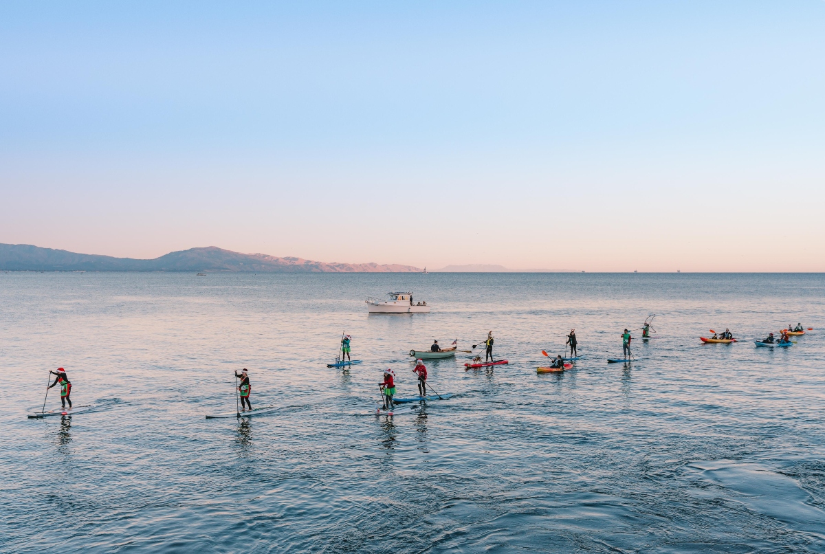 Multiple paddleboarders and kayakers are on calm ocean waters at sunset, with several participants wearing Santa hats and holiday-themed attire in red and green. A white motorboat is visible in the middle distance, and mountain ranges line the horizon under a pastel sky transitioning from blue to pink.