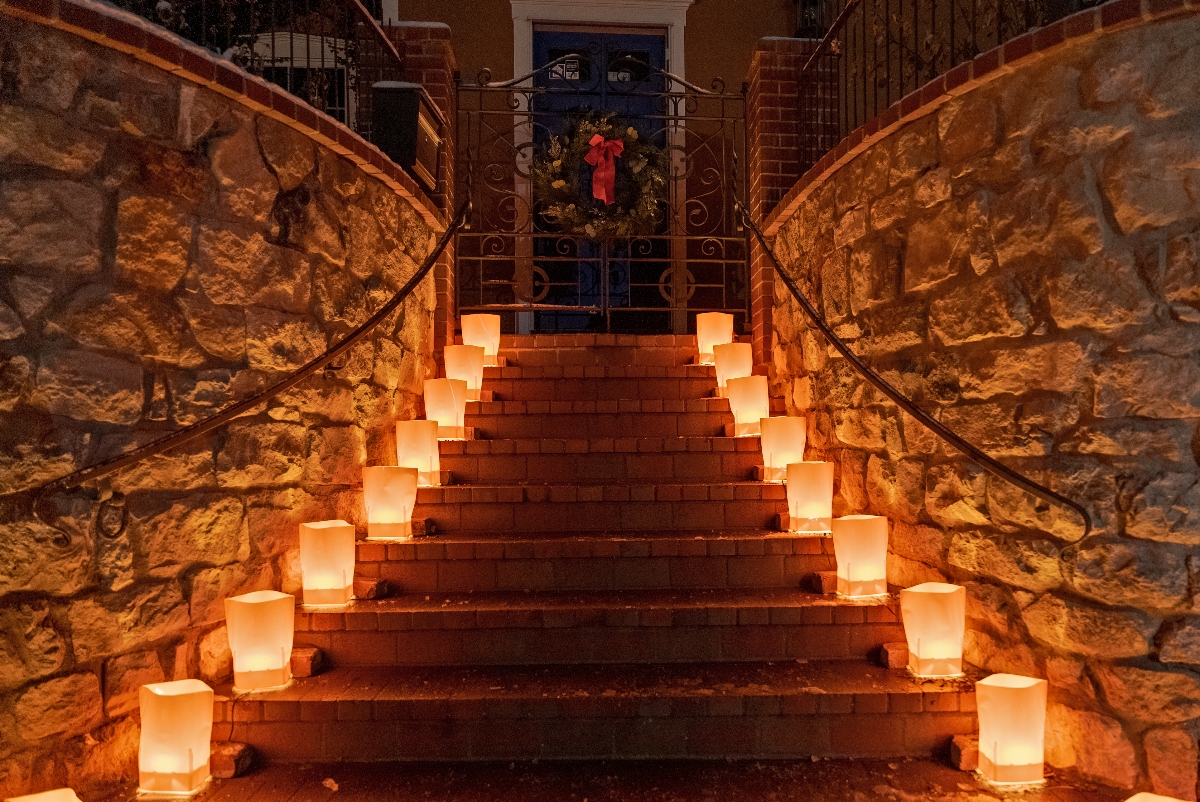 Brick stairs flanked by stone walls lead up to an ornate iron gate, with illuminated paper luminarias lining both sides of the stairway. A holiday wreath with a red ribbon hangs on the gate at the top of the stairs, and the warm glow from the luminarias creates golden lighting throughout the scene.