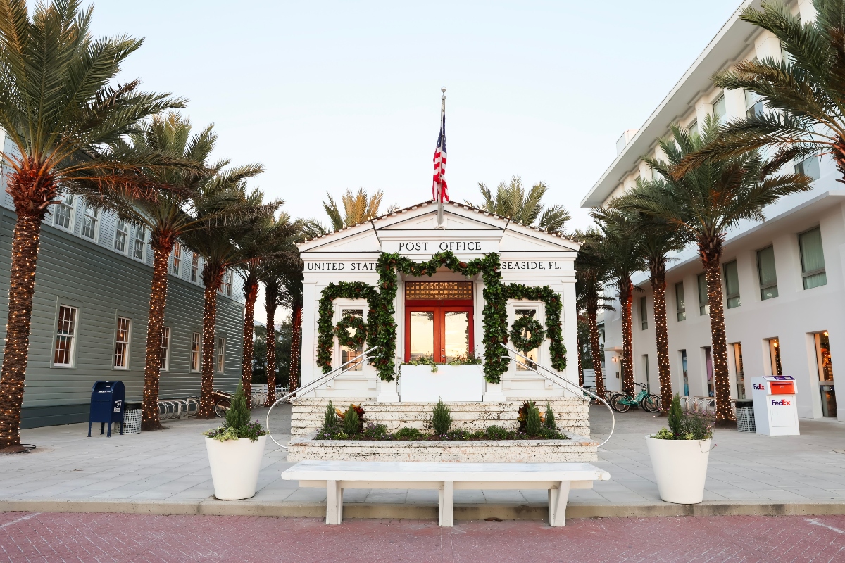 A white post office building in Seaside, Florida features a triangular pediment with an American flag on top, red double doors, and curved staircases leading to the entrance, all decorated with green garland and wreaths. Palm trees wrapped in string lights flank the building on both sides, with a white bench and planters in the foreground plaza area.