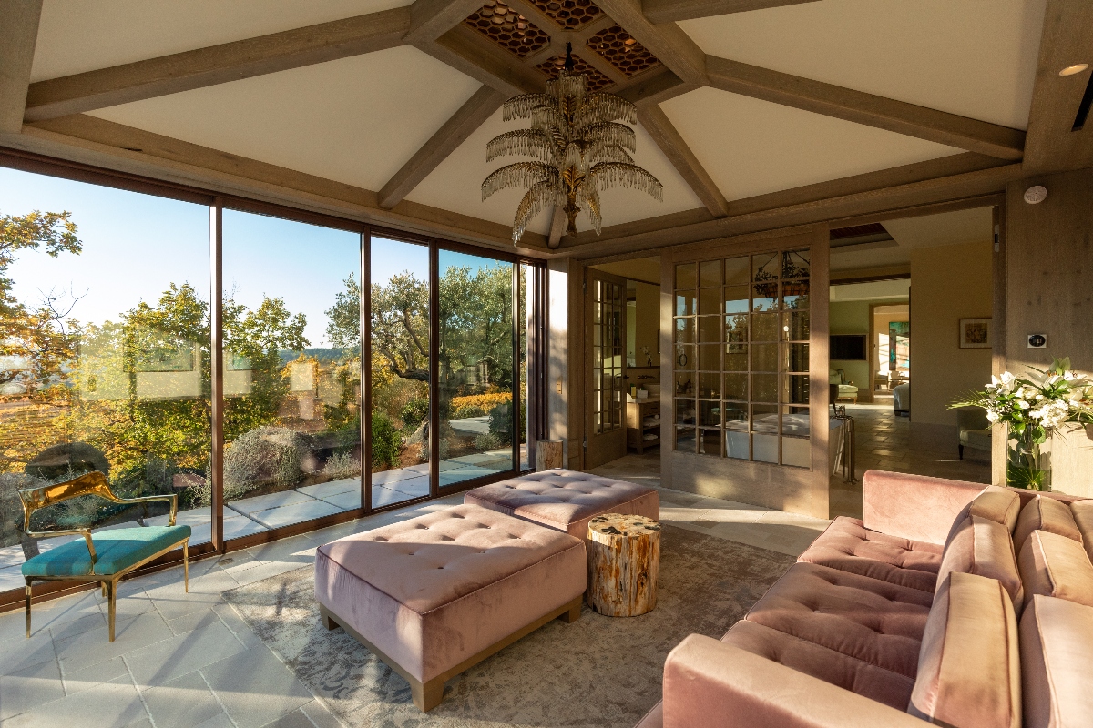 A spacious interior lounge features vaulted ceilings with exposed wooden beams, decorative tile inserts, and a multi-tiered crystal chandelier. The room is furnished with blush pink velvet tufted ottomans, a matching sectional sofa, and a natural wood stump side table on light stone tile flooring. Floor-to-ceiling windows and glass doors provide views of the surrounding garden and Provençal landscape, while French doors with divided glass panes connect to adjacent interior spaces.
