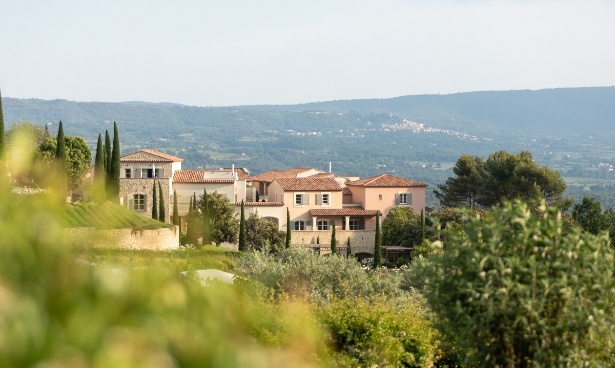 Mediterranean-style stone and stucco buildings with terracotta tile roofs sit on a hillside surrounded by cypress trees and lush vegetation in the Provence countryside. The property features multiple connected structures in cream, peach, and salmon tones with blue shutters. Rolling hills with scattered buildings and a hilltop village extend across the background beneath a hazy sky.