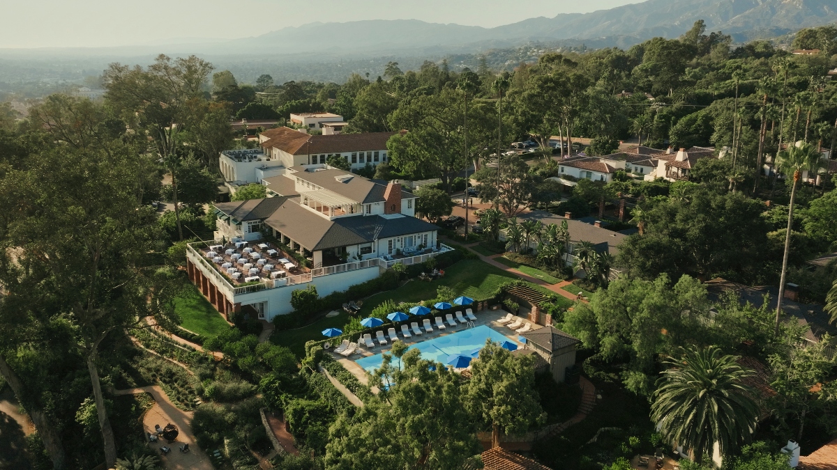 An aerial view captures the sun-drenched elegance of a hillside Spanish colonial resort, where white-washed buildings with terracotta roofs nestle among towering palms and verdant gardens overlooking the Santa Barbara coastline and misty mountain ranges beyond. The property's jewel-like pool, dotted with vibrant blue umbrellas, anchors multiple levels of manicured terraces that cascade down the lush, tree-lined hillside.