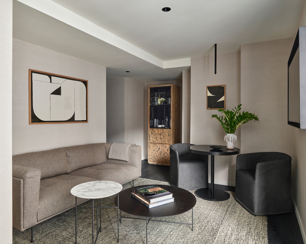 A sophisticated study in neutral minimalism unfolds through this contemporary suite, where textured linen-wrapped walls provide an elegant backdrop for a curvaceous taupe sofa, sculptural white marble and black lacquer nesting tables, and bold geometric artwork framed in warm wood. The refined space flows seamlessly into an intimate dining area anchored by sleek black furnishings and a statement bar cabinet of exotic burl wood, its glass shelves gleaming with crystal and greenery adding a fresh touch of life to the monochromatic palette.