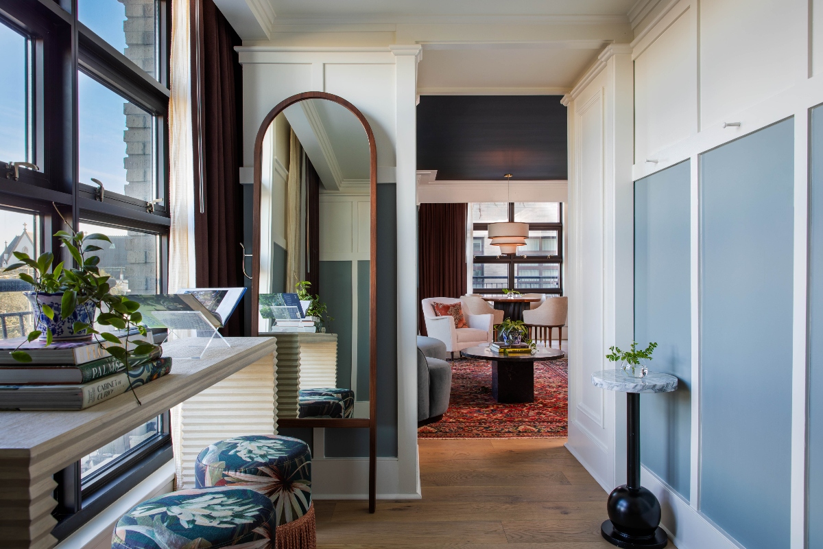 A sun-filled hotel suite unfolds through classic architectural details, where a stately arched mirror anchors a cozy window-side perch styled with blue ikat ottomans and curated coffee table books, while floor-to-ceiling windows frame urban rooftop views beyond flowing curtains. The sophisticated space flows seamlessly into an elegant sitting room visible through white-paneled doorways, where plush blush velvet chairs and a dramatic abstract painting create an inviting backdrop for conversation over a richly patterned Persian rug.