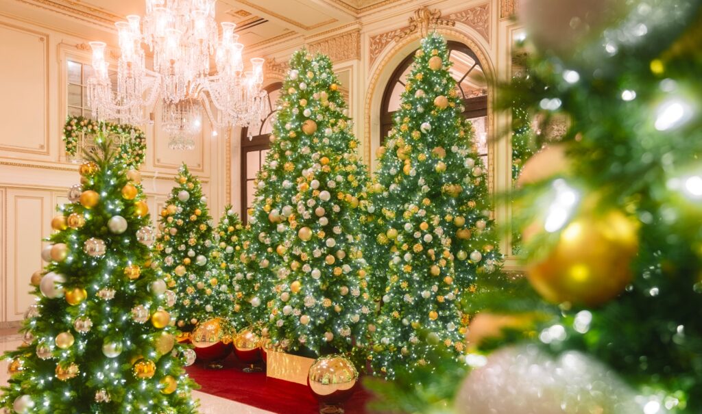 Multiple decorated Christmas trees adorned with gold and silver ornaments stand in an ornate hotel lobby featuring cream-colored walls with gilded molding, arched doorways, and crystal chandeliers. A red carpet runner extends between the illuminated trees, with wrapped gift boxes displayed near the center tree.