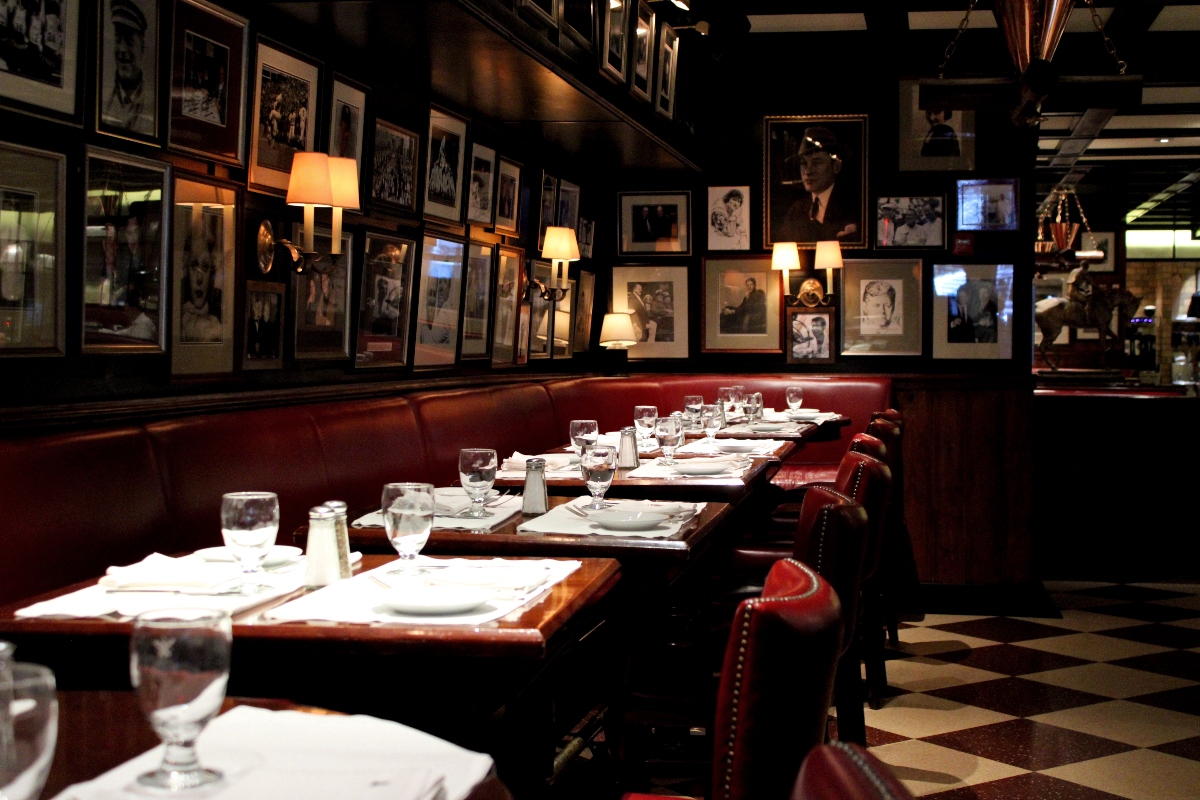Interior of a classic steakhouse dining room featuring dark walls lined with black and white framed photographs, illuminated by warm wall sconces. Red leather banquette seating runs along the wall beside a row of wooden tables set with white linens, water glasses, and place settings on a black and white checkered floor.
