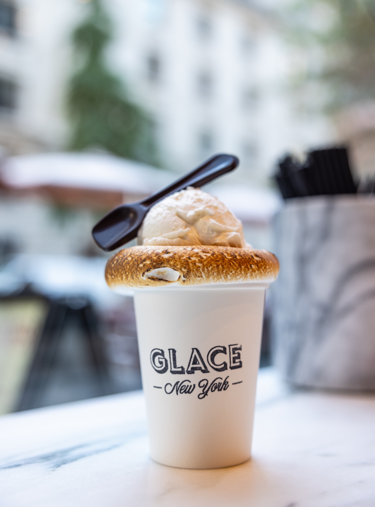 A white paper cup branded "GLACE New York" holds a scoop of vanilla ice cream topped with a toasted marshmallow that shows golden-brown caramelization. A small black spoon rests on top of the dessert, and the cup sits on a white surface with a blurred cafe interior visible in the background.