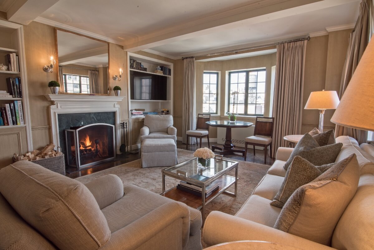 A living room with beige and taupe furnishings features a white-manteled fireplace with an active fire, built-in bookshelves, and a flat-screen television mounted in cabinetry. The space includes upholstered seating arranged around a glass coffee table, a round wooden dining table near tall windows with floor-length curtains, and warm lighting from wall sconces and table lamps.