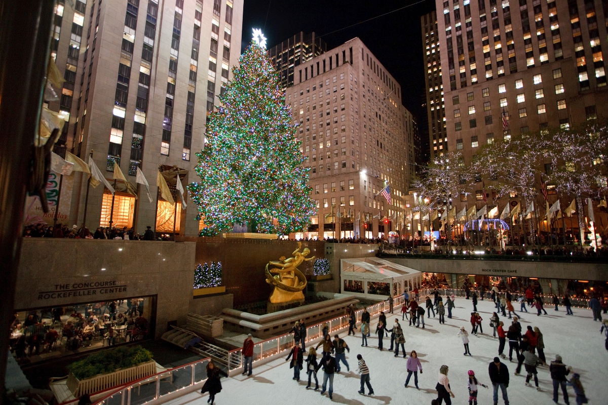 The Rockefeller Center Christmas tree stands illuminated with thousands of multicolored lights and a white star topper, towering over the ice skating rink below where dozens of people skate. The golden Prometheus statue is positioned between the tree and the rink, with Art Deco buildings rising on all sides, their windows glowing against the night sky. Holiday decorations line the plaza level, and crowds of visitors fill the concourse area beneath the rink.