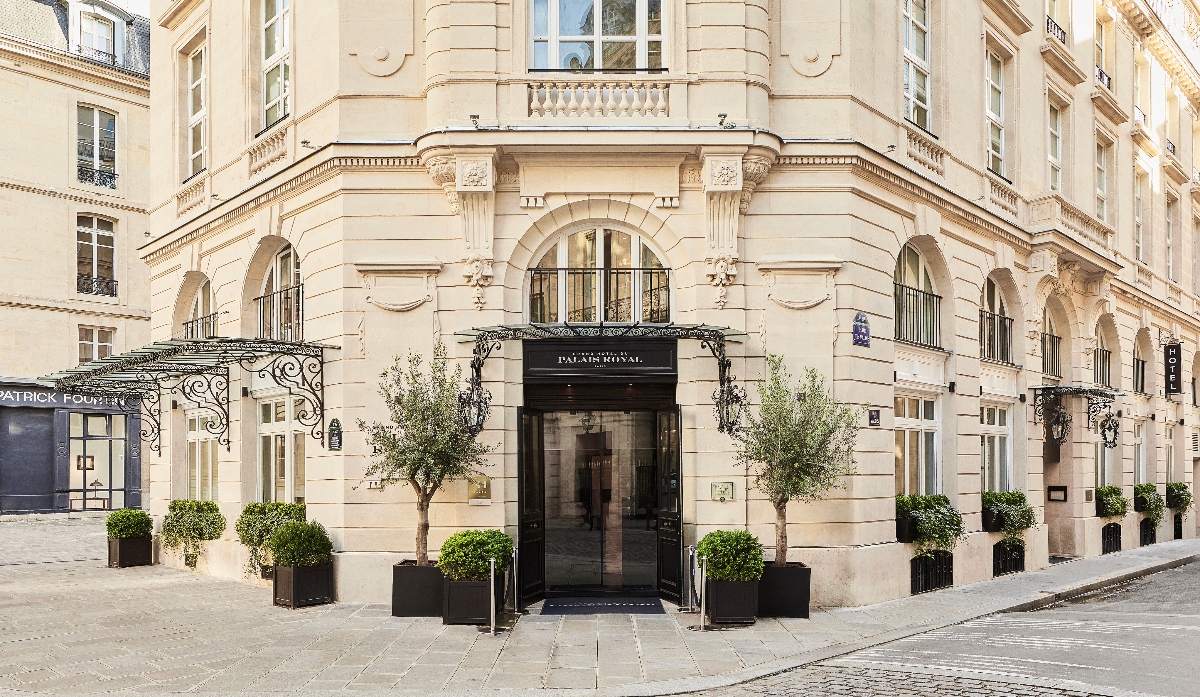 Hotel exterior featuring a cream-colored stone building with classical Haussmann-style architecture, including arched windows, decorative balustrades, ornamental columns, and carved stonework details across multiple floors. The ground-level entrance has black glass doors beneath a black canopy with ornate wrought-iron scrollwork, flanked by large planters containing trimmed topiary and small trees. The building sits on a cobblestone street with adjacent storefronts and residential buildings visible on either side.