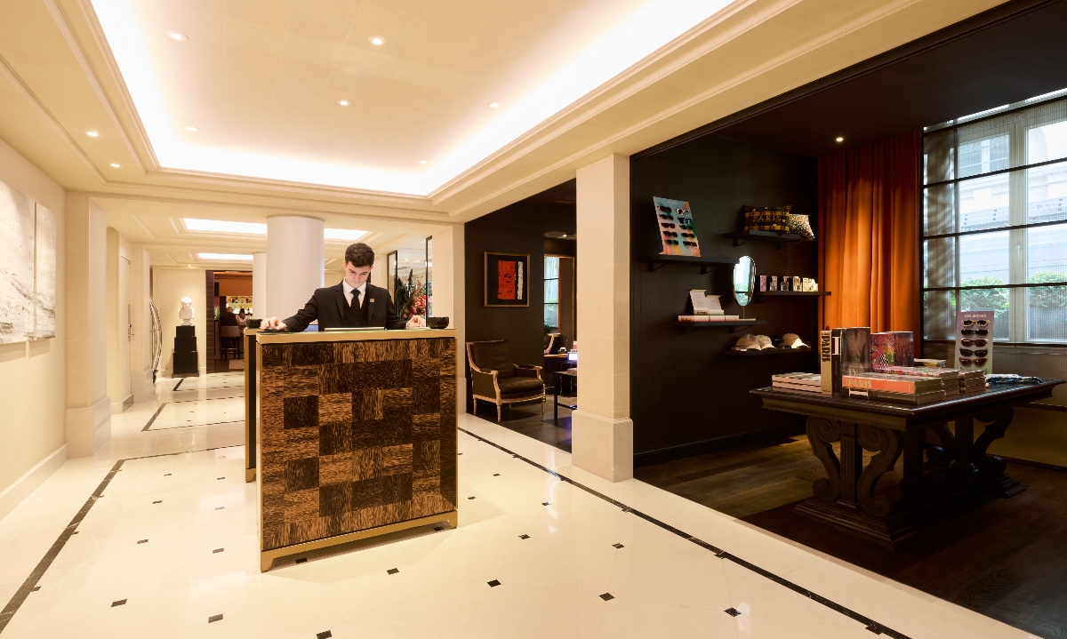 Hotel lobby featuring white marble flooring with dark diamond inlays and a concierge desk with wood-grain paneling in the foreground, where a staff member in formal attire stands at attention. The space includes cream walls with decorative crown molding, recessed ceiling lighting, and an adjacent sitting area with dark walls, built-in shelving displaying books and decorative objects, and upholstered seating near tall windows with sheer drapery.