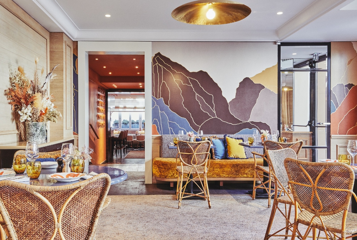 Restaurant interior featuring woven rattan dining chairs arranged around tables set with glassware and floral arrangements, beneath a large brass dome pendant light fixture. The space includes a gold velvet banquette with blue and yellow accent pillows positioned against a wall-mounted artistic mural depicting stylized mountains in brown and purple tones with white linear detailing. An archway with terracotta-painted ceiling provides a view into an adjacent dining room, while glass partition doors separate other areas, with light wood paneling and gray carpeting throughout.