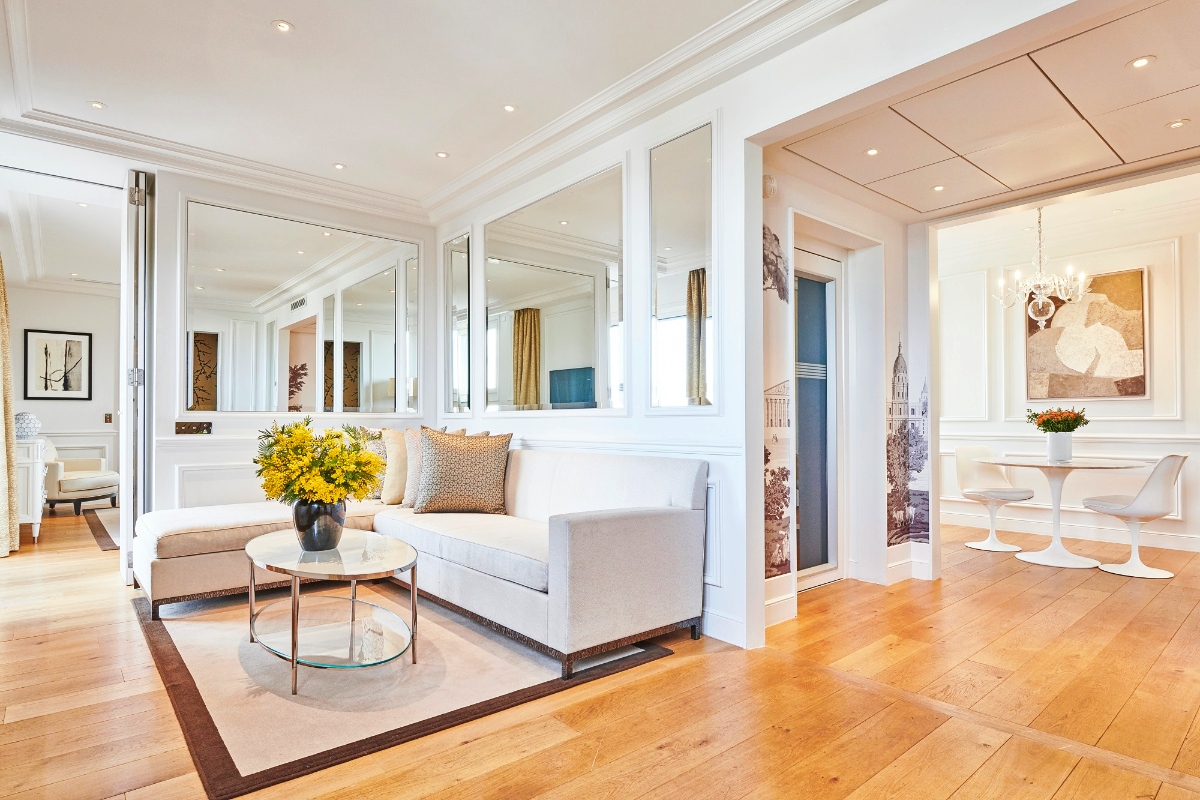 Hotel suite interior featuring light wood plank flooring throughout an open-plan living and dining space with white walls, decorative molding, and recessed lighting. The living area includes a cream-colored sofa with decorative pillows arranged around a round glass-top coffee table displaying yellow flowers, positioned on a light area rug with dark border trim. Adjacent spaces include a dining area with a white pedestal table and matching chairs, plus additional rooms visible through doorways, with wall-mounted mirrors and framed artwork throughout.